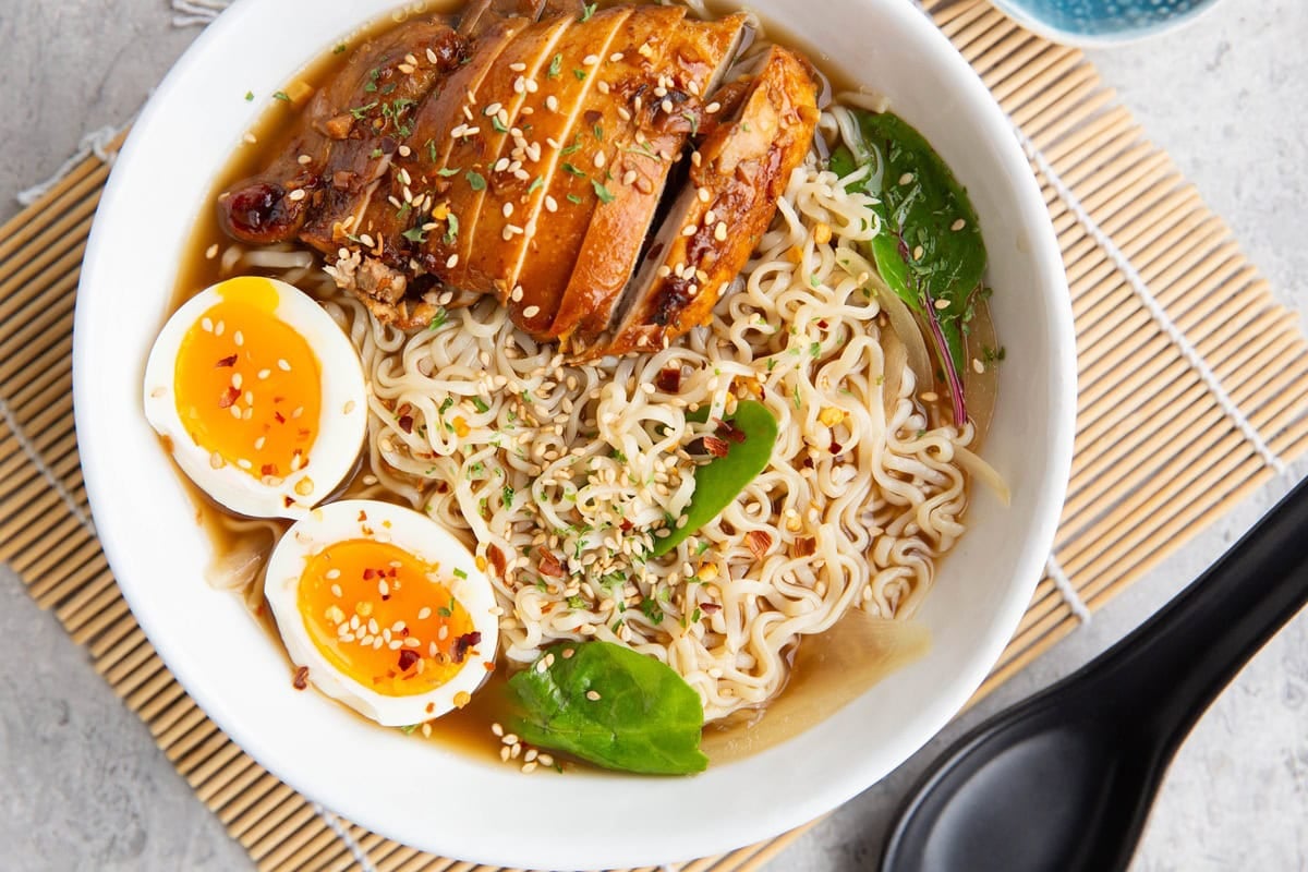 Ramen in a white bowl with broth, soft boiled eggs cut in half, and sliced chicken. A black Chinese soup spoon to the side. Ready to eat.