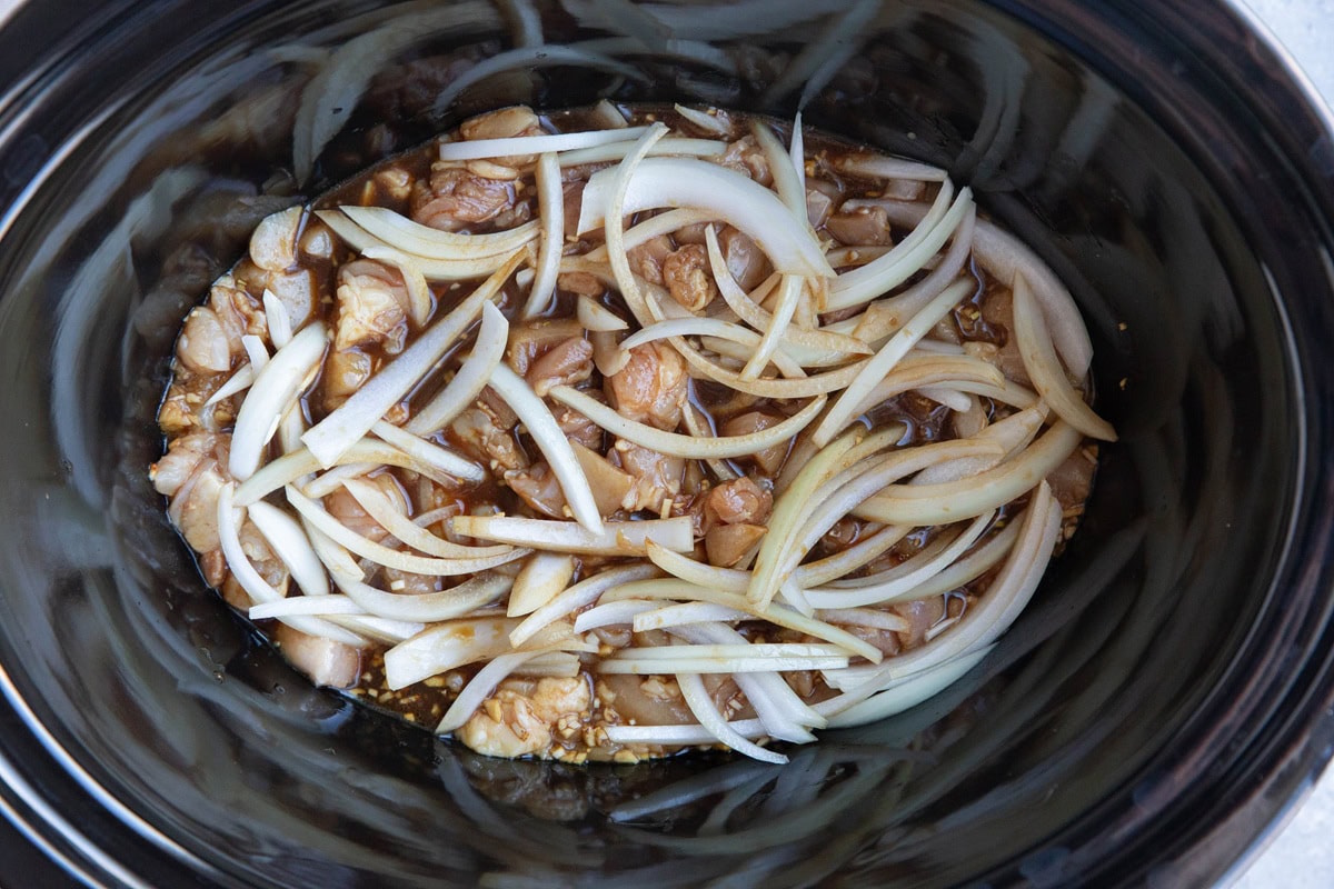 Crock pot full of teriyaki sauce with chopped chicken thighs and sliced onions.