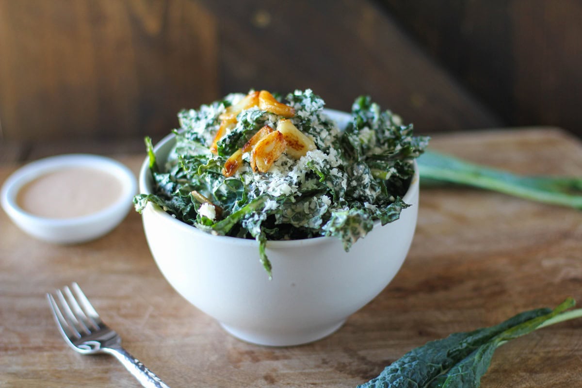 Large white bowl of kale Caesar salad with a small bowl of dressing in the background and fresh kale leaves to the side.