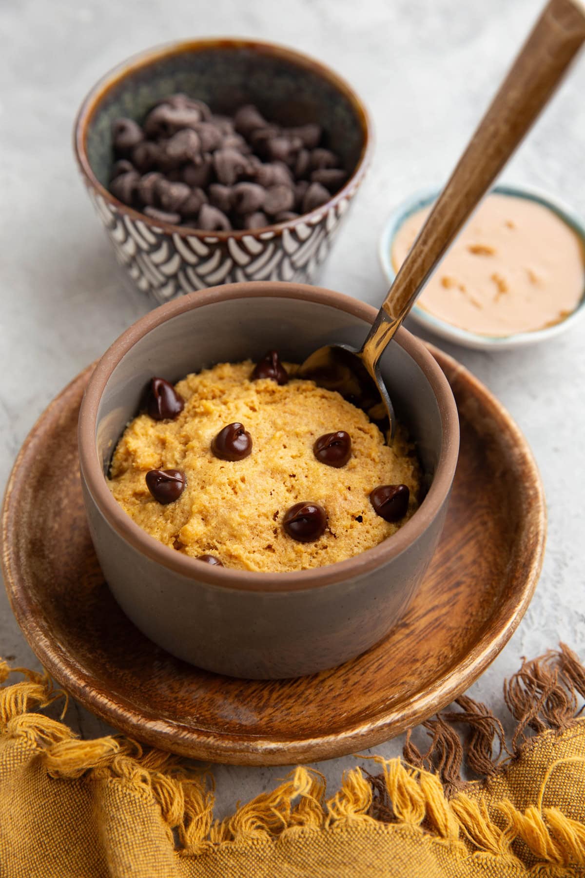Peanut butter cake in a ramekin sitting on a wooden plate with a small bowl of peanut butter in the background and a bowl of chocolate chips.