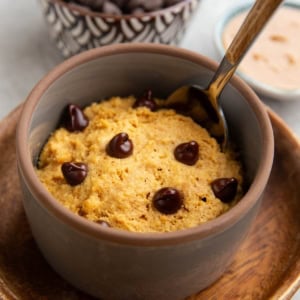 Peanut butter mug cake in a grey ramekin on a small wooden plate with a bowl of chocolate chips and peanut butter in the background and a spoon inside of the mug cake, ready to serve.