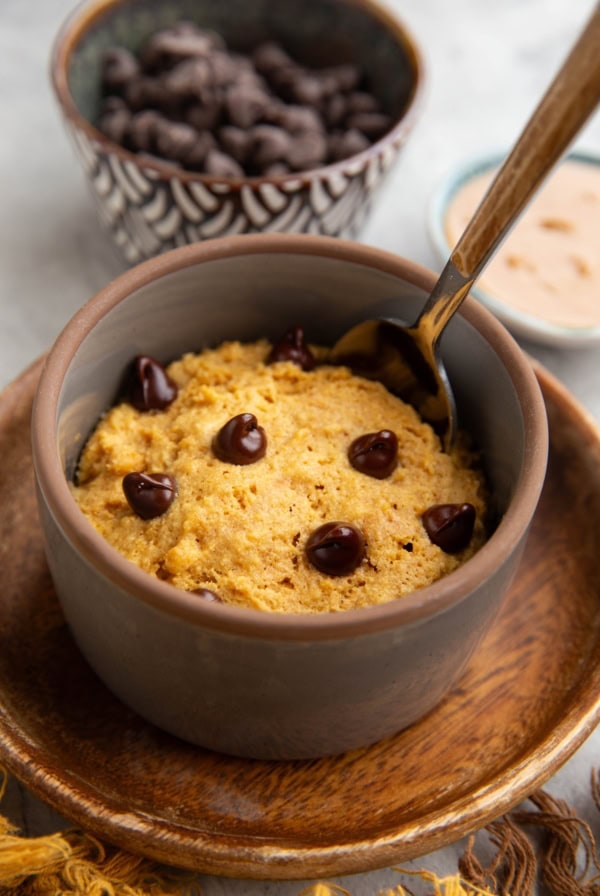 Peanut butter mug cake in a grey ramekin on a small wooden plate with a bowl of chocolate chips and peanut butter in the background and a spoon inside of the mug cake, ready to serve.