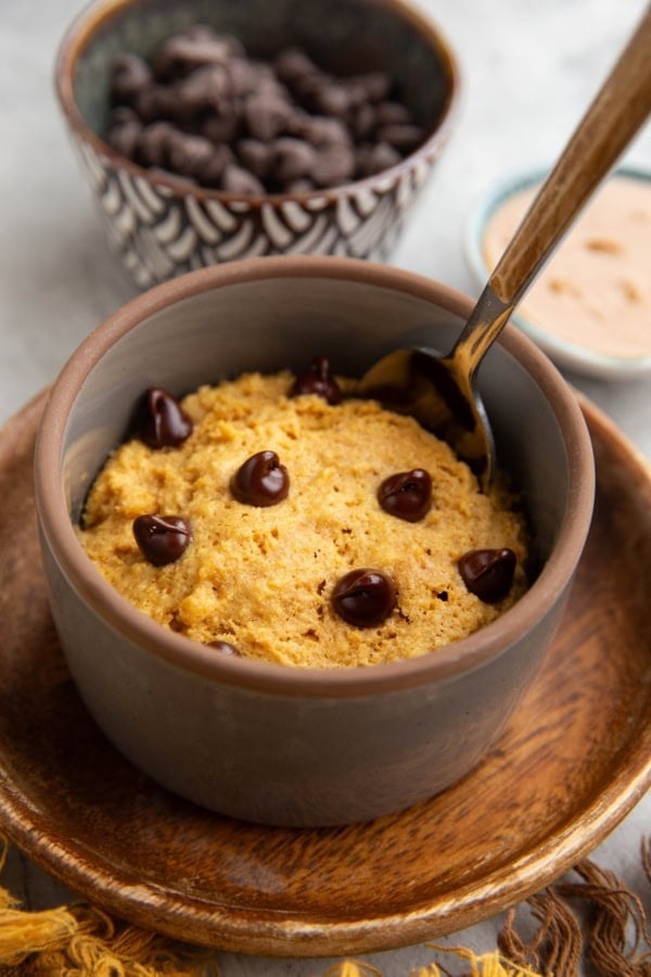 Peanut butter mug cake in a grey ramekin on a small wooden plate with a bowl of chocolate chips and peanut butter in the background and a spoon inside of the mug cake, ready to serve.