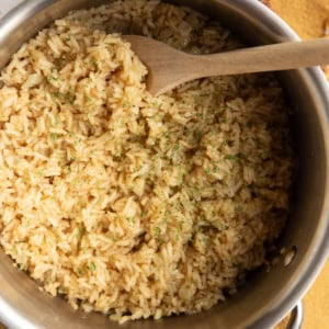 Chicken rice in a stainless steel pot sprinkled with dried parsley and a wooden spoon scooping some of it out. A bowl of dried parsley to the side and a golden napkin sits underneath the pot.