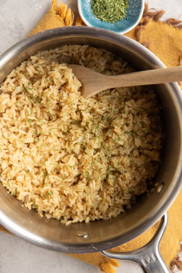 Chicken rice in a stainless steel pot sprinkled with dried parsley and a wooden spoon scooping some of it out. A bowl of dried parsley to the side and a golden napkin sits underneath the pot.