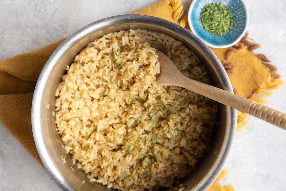 Chicken rice in a stainless steel saucepan with a wooden spoon inside the pot scooping some of the rice. A small bowl of dried parsley sits to the side.