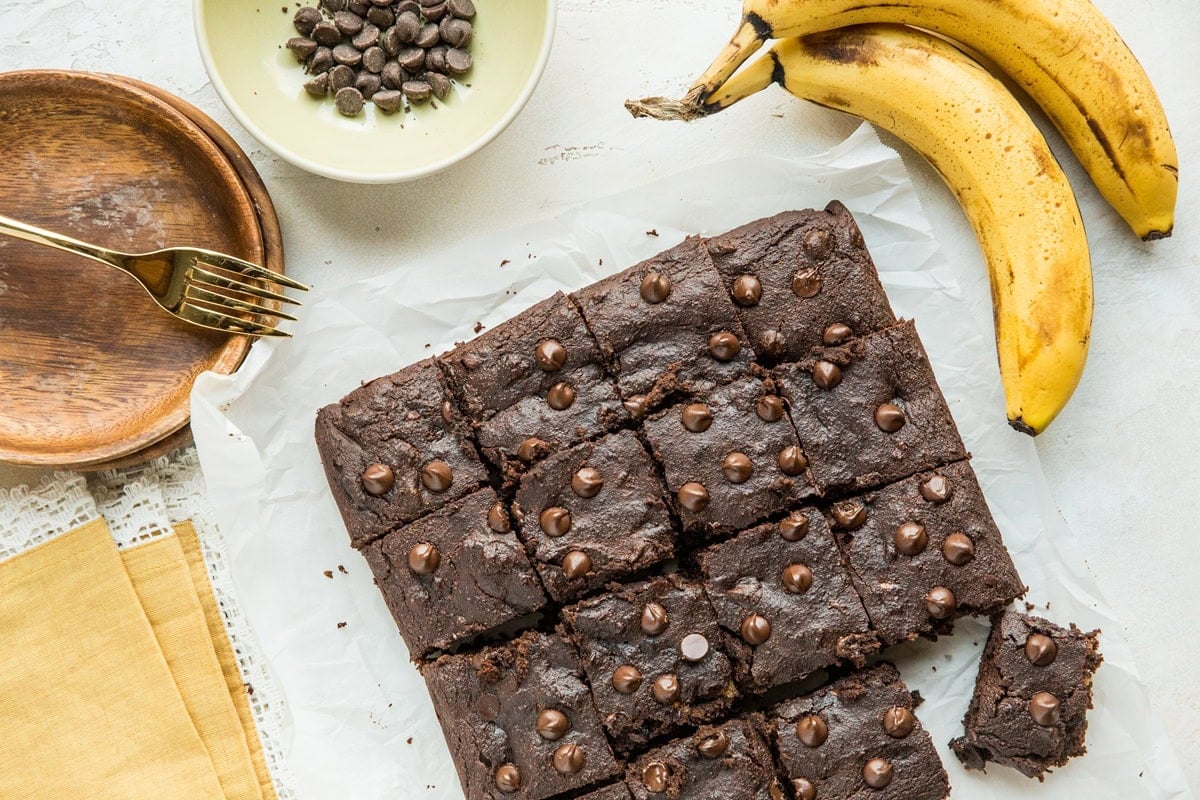 Chocolate cake cut into squares on a sheet of paper with ripe bananas and a bowl of chocolate chips to the side.