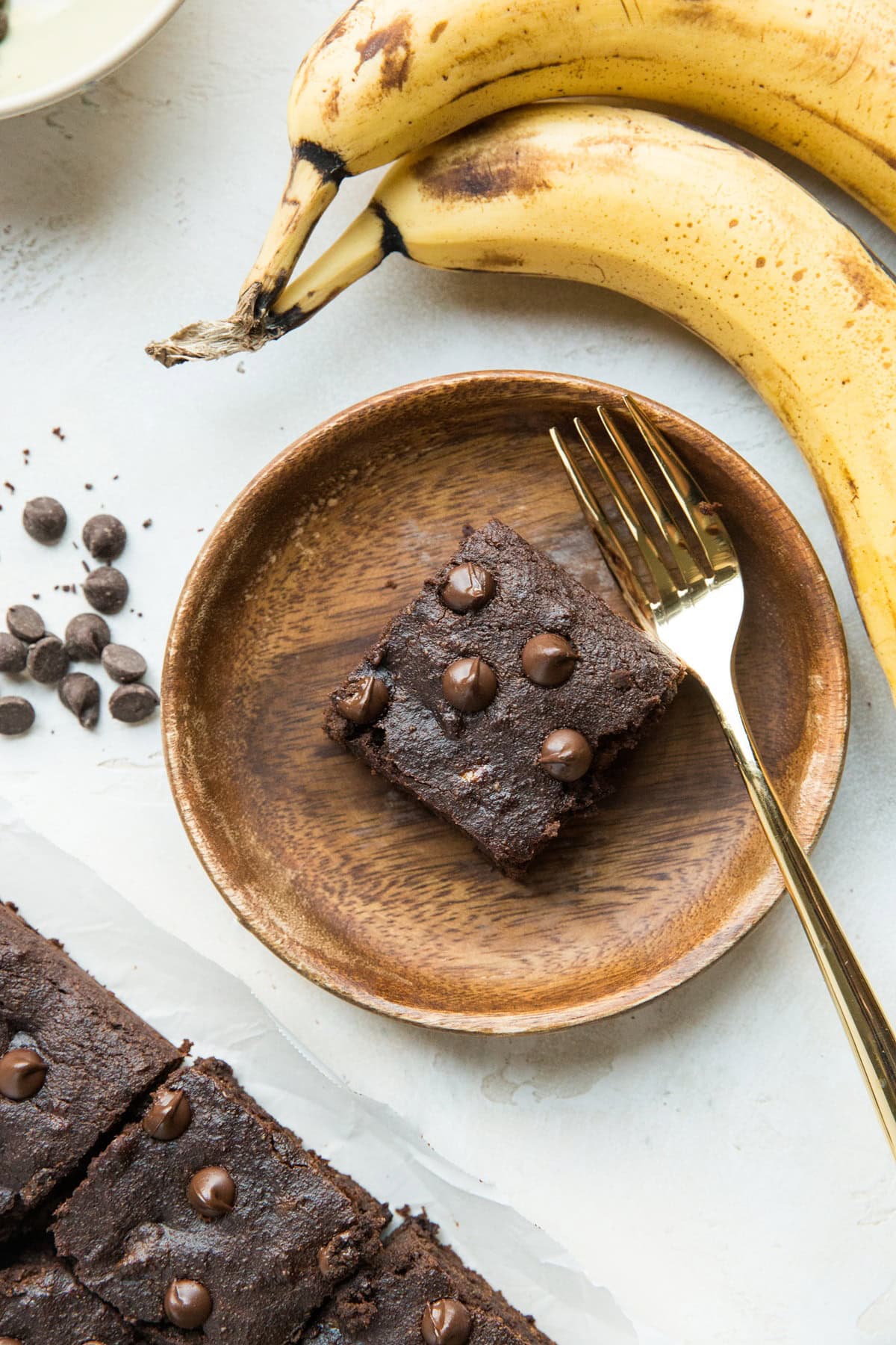 Wooden plate with a slice of chocolate cake and a gold fork to the side for eating. Ripe bananas to the side and stray chocolate chips.