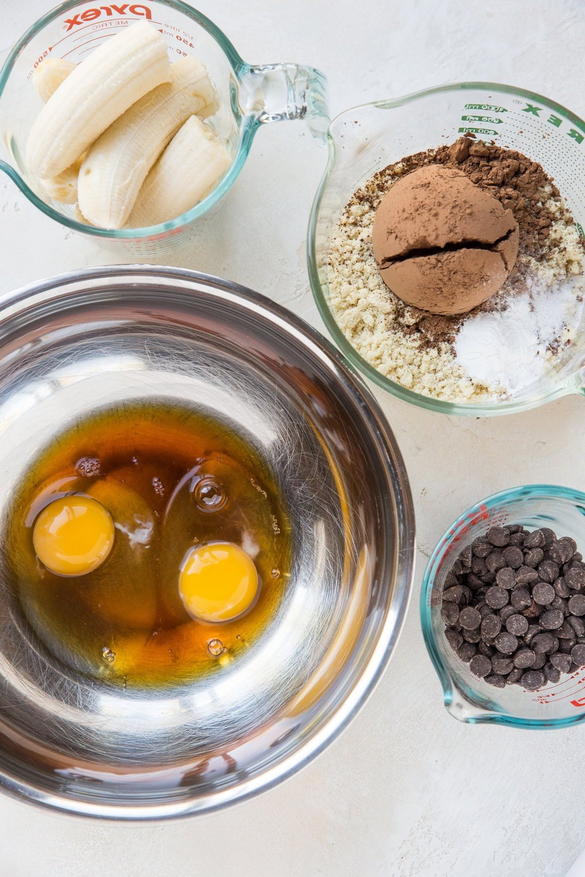 Stainless steel bowl full of two eggs, pure maple syrup and oil, a large measuring cup with almond flour, cocoa powder, and baking powder, a measuring cup full of ripe bananas and a measuring cup of chocolate chips. These are the ingredients for our cake.