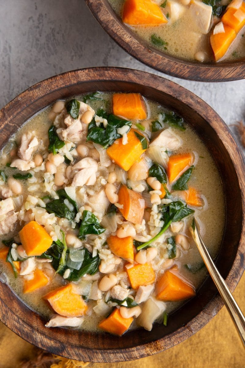 Two wooden bowls full of chicken and sweet potato soup with a gold spoon and a gold napkin to the side.