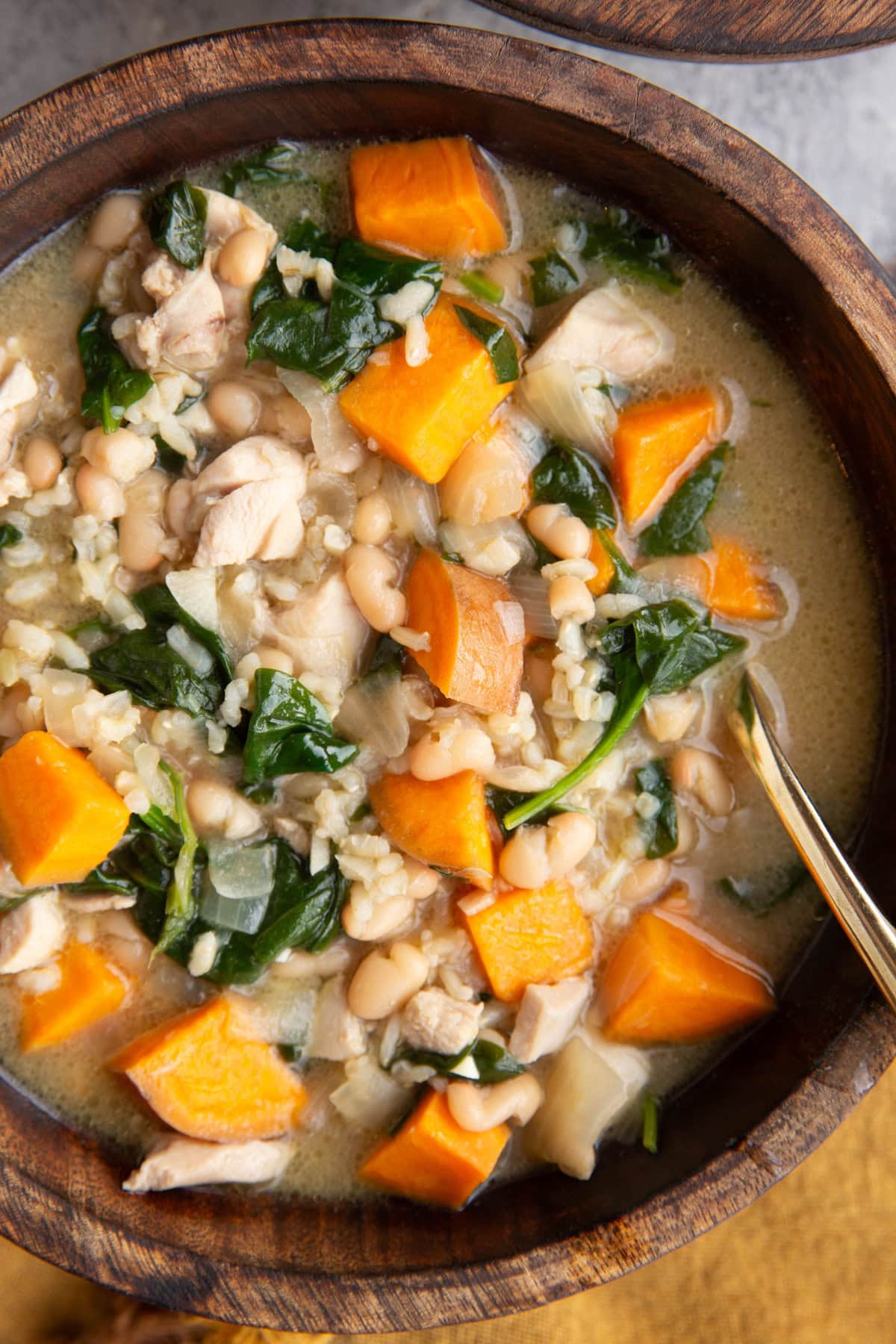 Large wooden bowl full of creamy chicken and sweet potato soup with spinach and white beans and brown rice. A napkin to the side and a gold spoon inside of the soup for eating.
