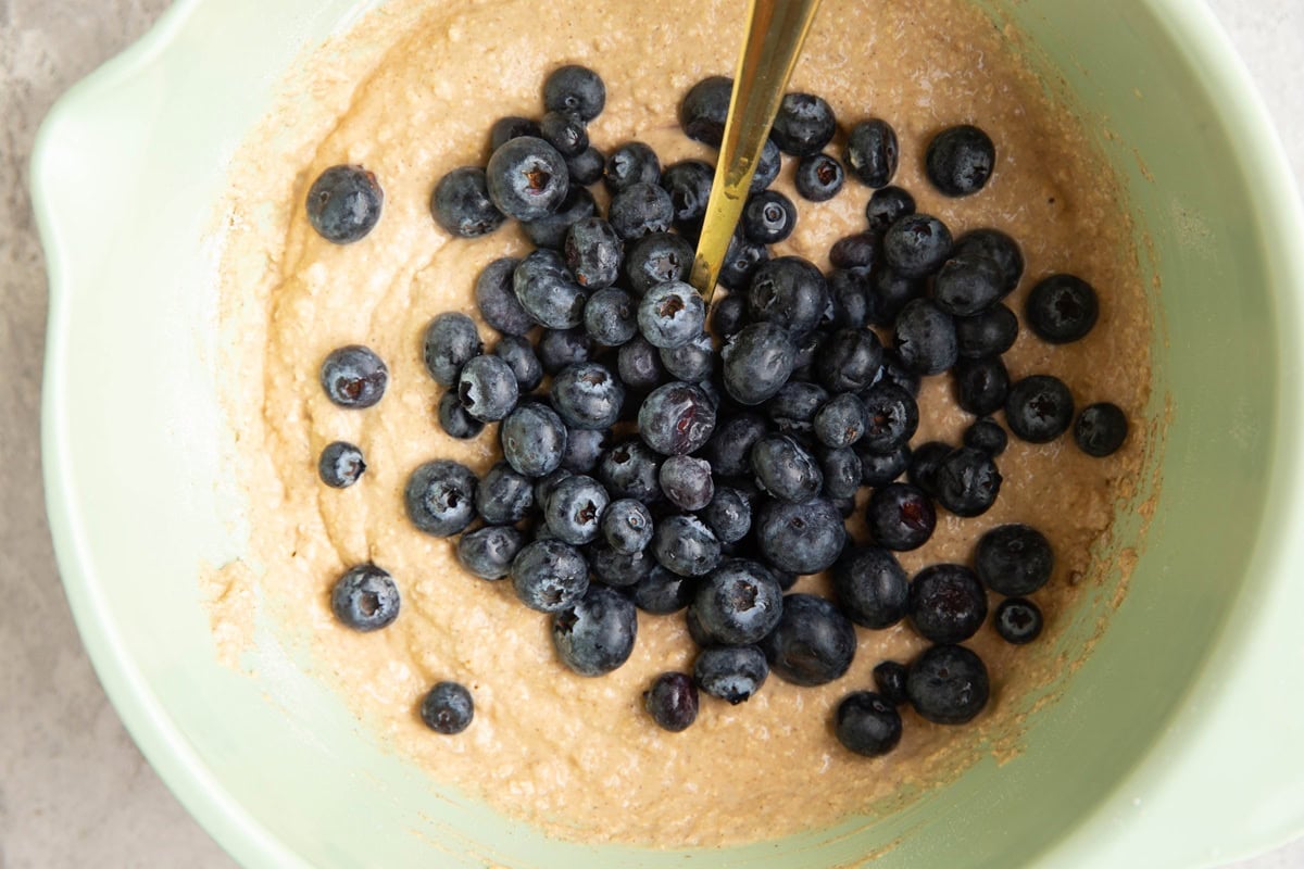 Mixing bowl with oatmeal bread mixture on the bottom and fresh blueberries on top, ready to be mixed into the batter.