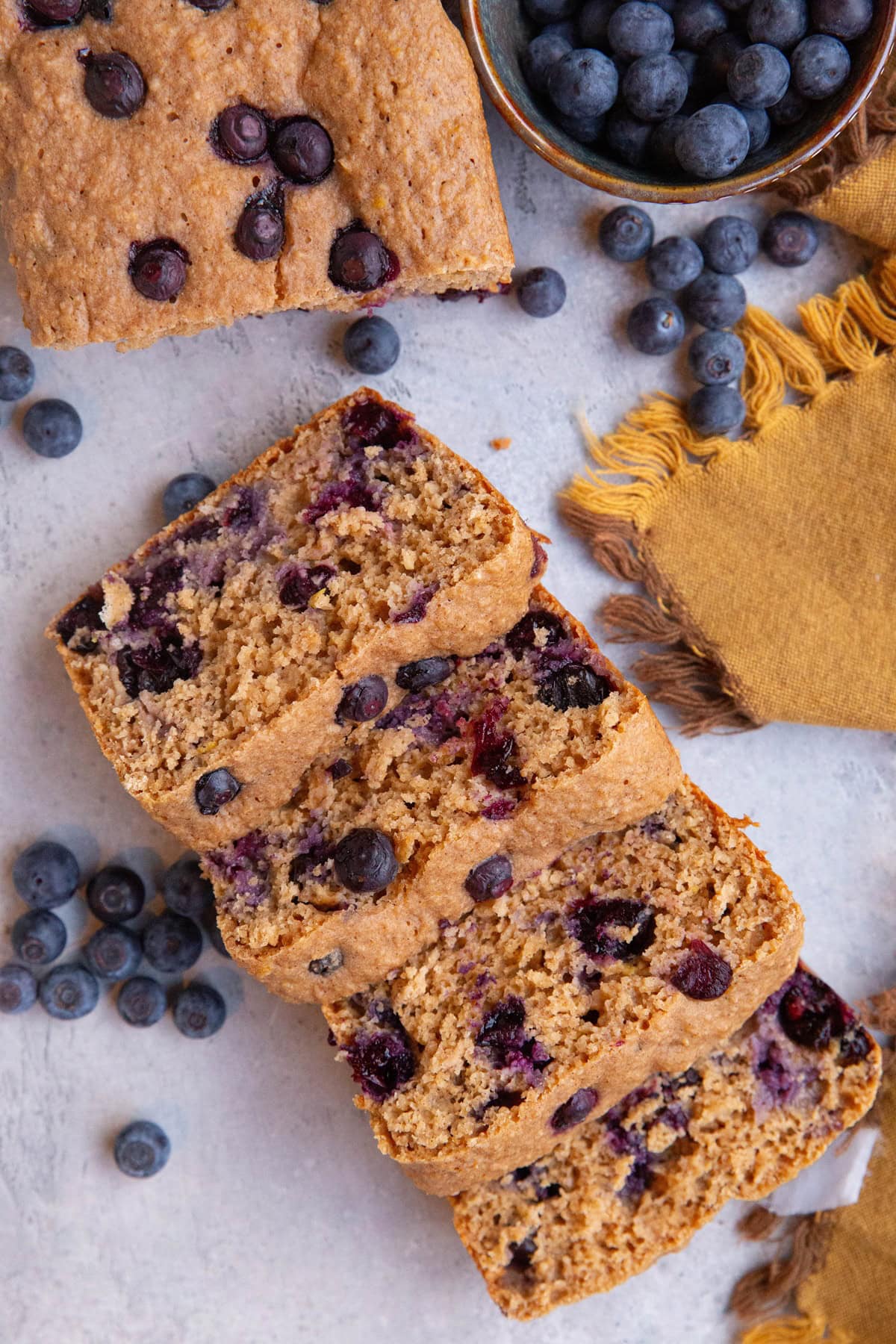 Loaf of blueberry bread cut into slices with a bowl of fresh blueberries to the side and a golden napkin.