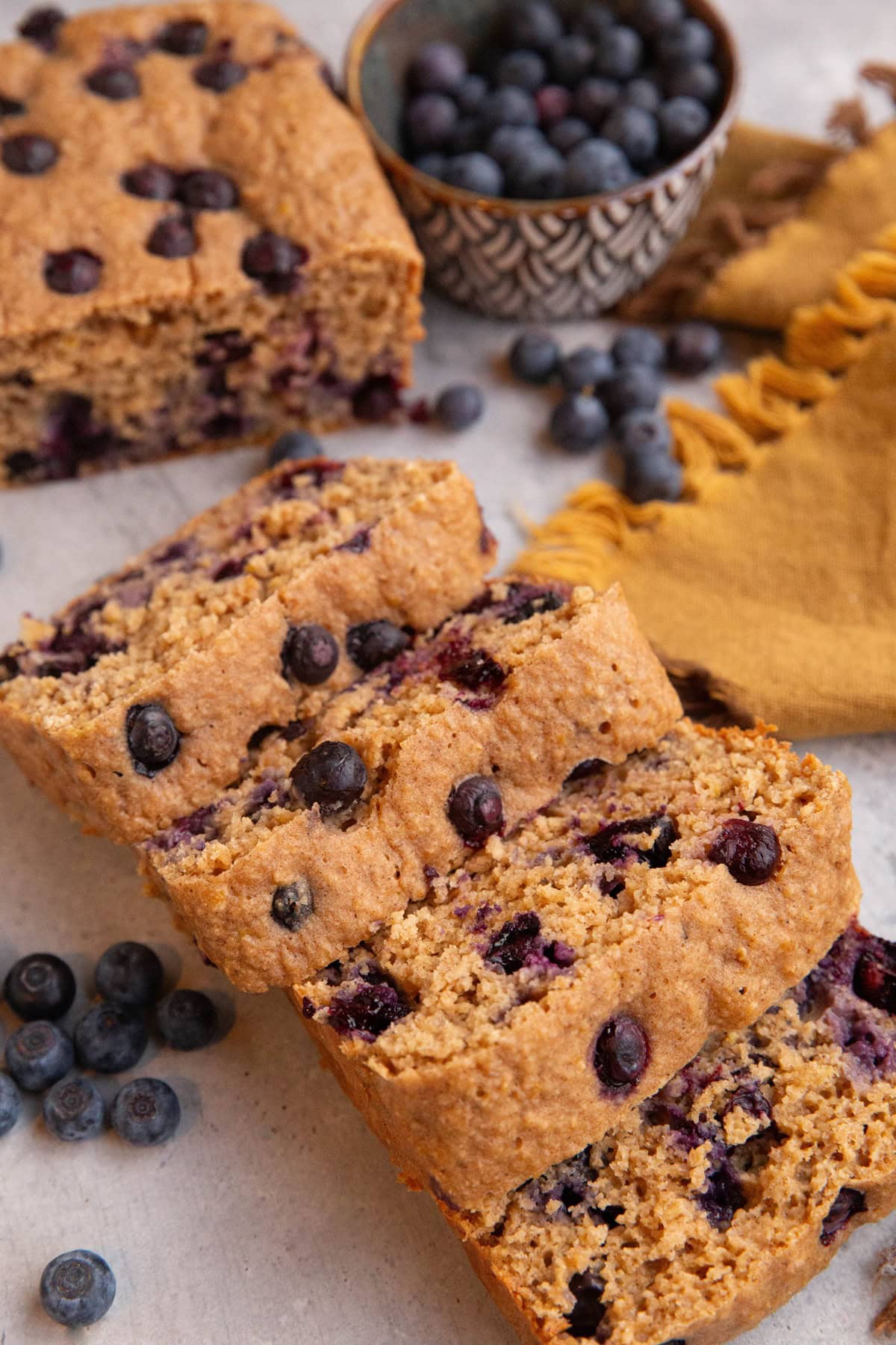 Loaf of blueberry oatmeal yogurt bread cut into slices with a bowl of fresh blueberries in the background.