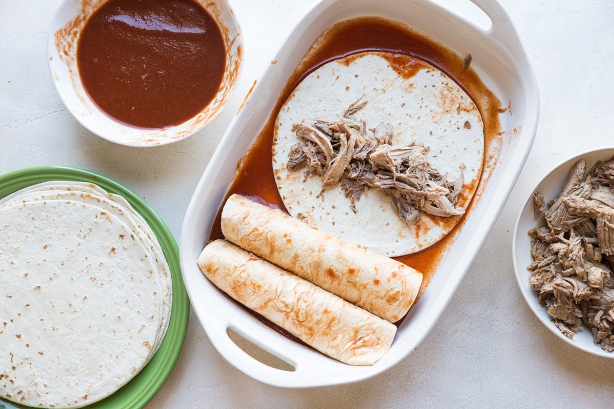 Casserole dish with two enchiladas made, one in the process of being made with shredded pork in the center, and bowls of enchilada sauce, pork, and tortillas to the side to make pork enchiladas.