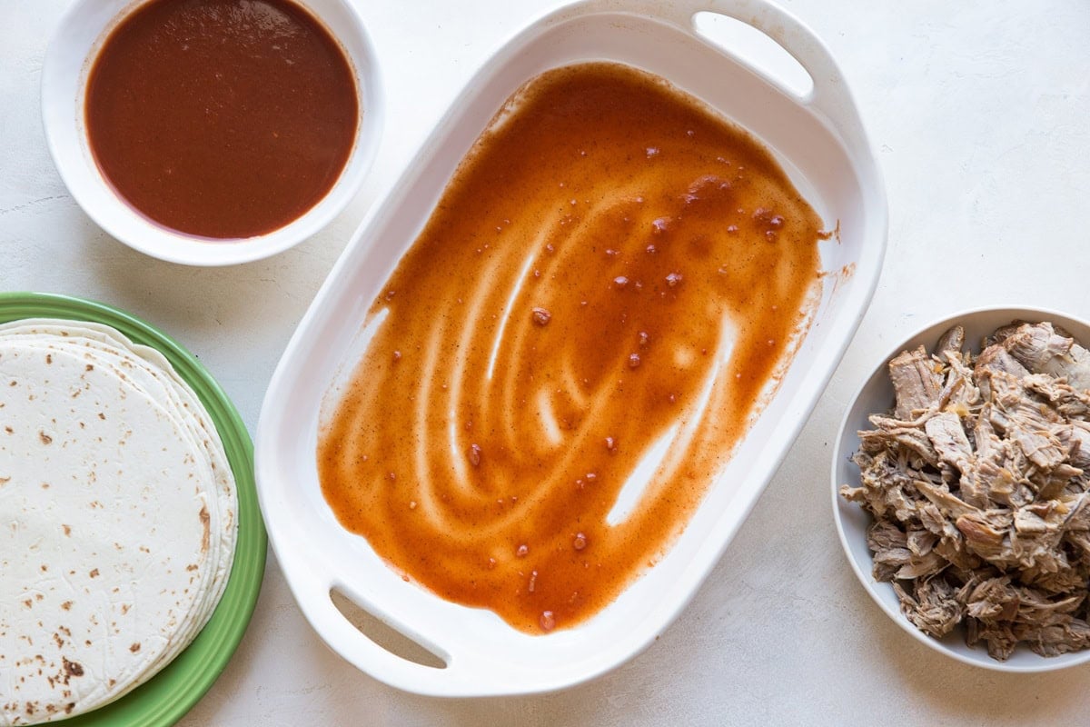 Casserole dish with enchilada sauce spread on the bottom with another bowl of enchilada sauce to the side, a bowl of shredded pork, and a plate full of flour tortillas.