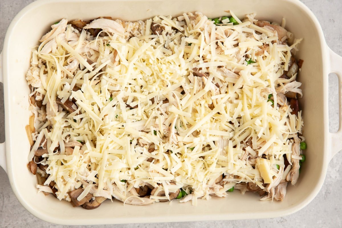 large casserole dish full of rotisserie chicken, vegetables, rice, and sprinkled with grated cheese on top, ready to go into the oven.