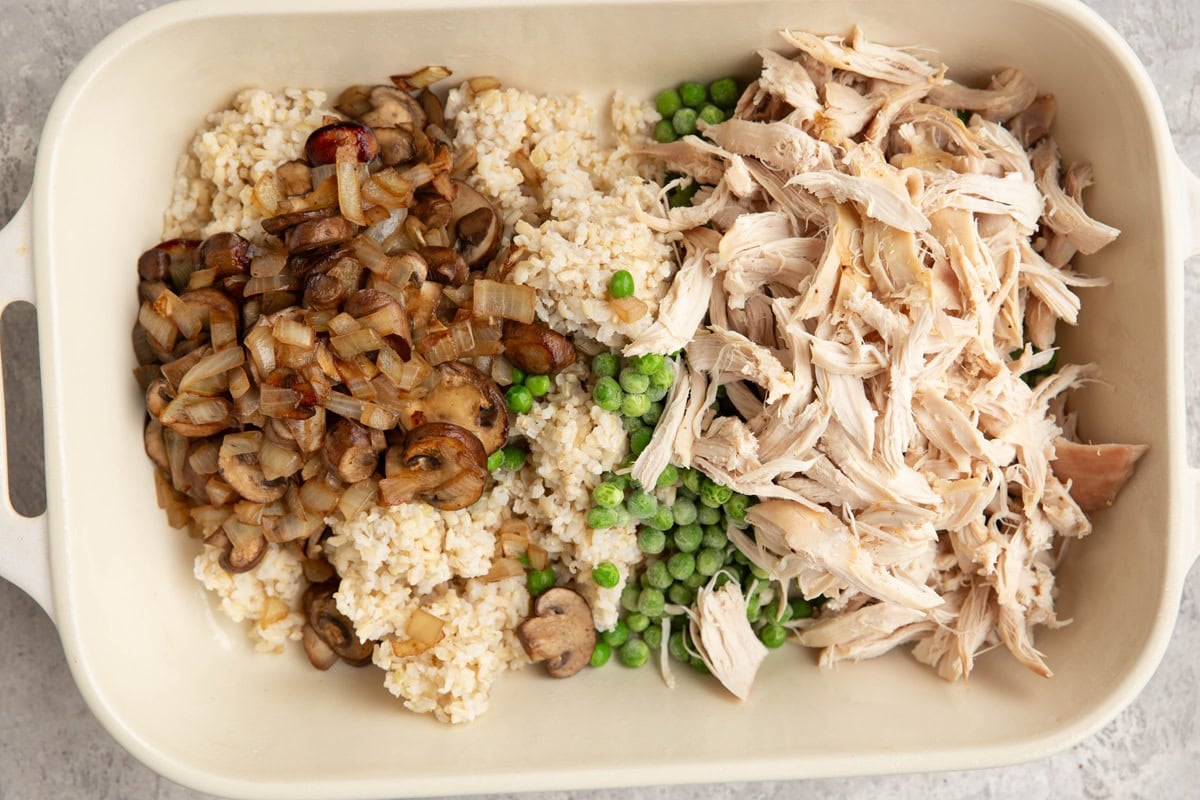large casserole dish full of shredded chicken, cooked brown rice, sauteed onion and mushrooms, and peas.
