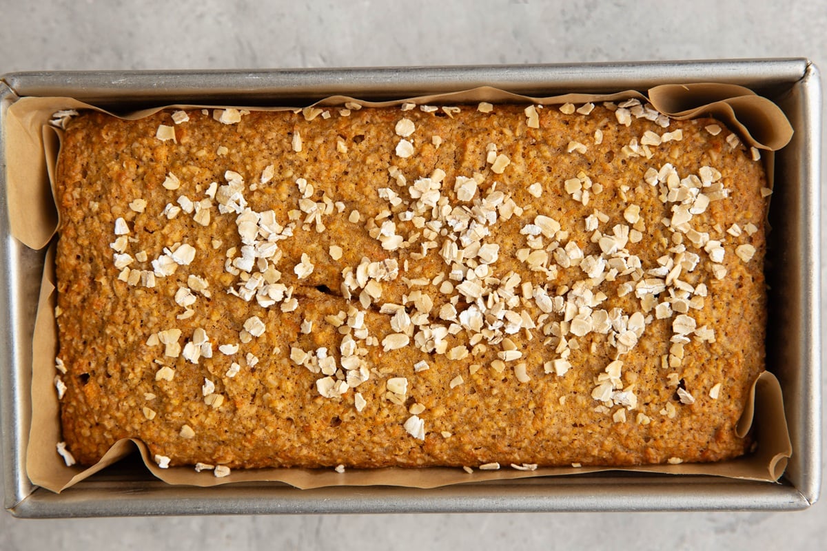Loaf pan lined with parchment paper with sweet potato bread inside, sprinkled with oats, fresh out of the oven.