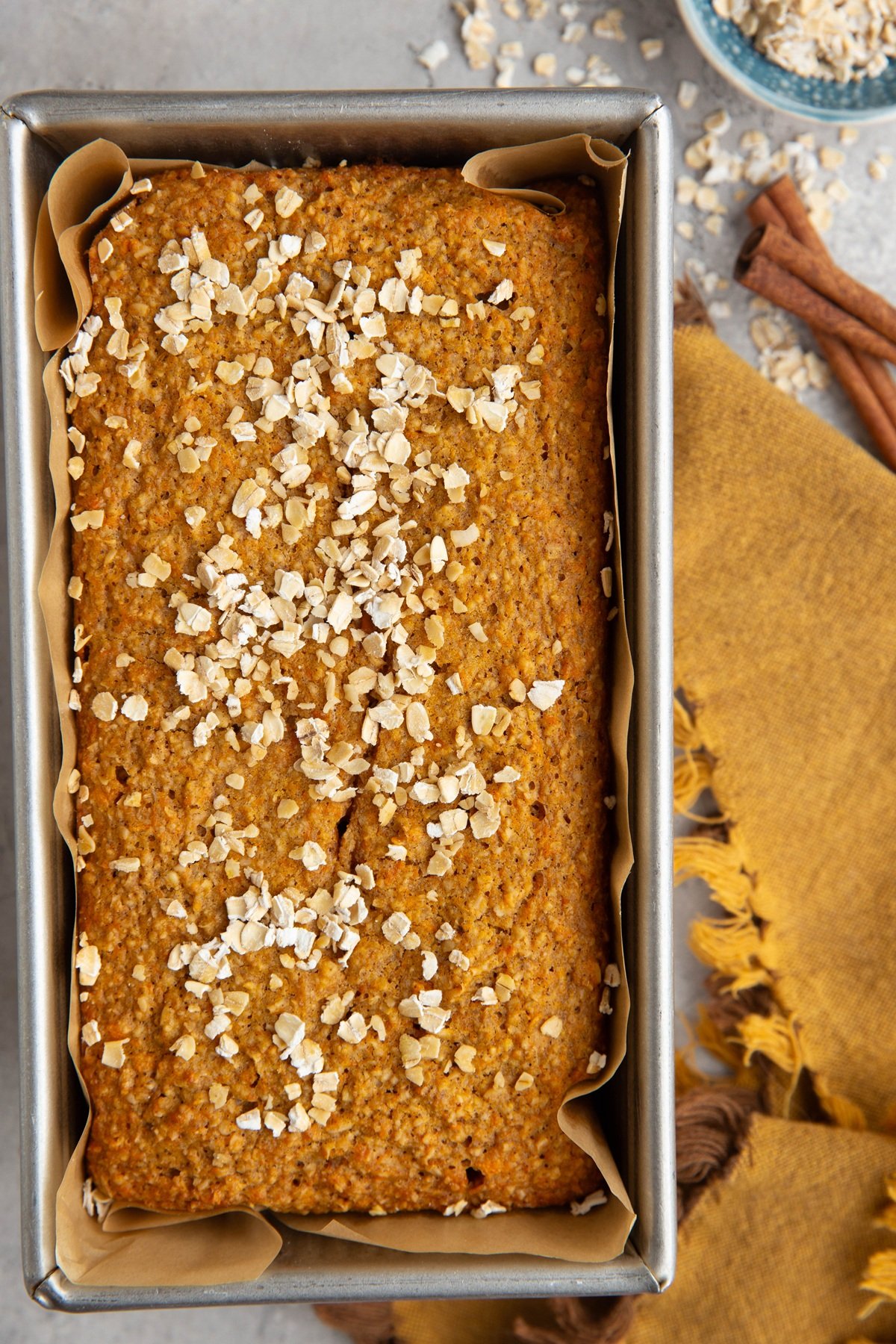 Sweet potato bread in a loaf pan with oats sprinkled on top, oats scattered to the side next to cinnamon sticks and a golden napkin.