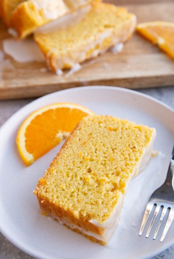 Slice of orange pound cake on a white plate with the rest of the pound cake cut into slices in the background. Ready to serve.