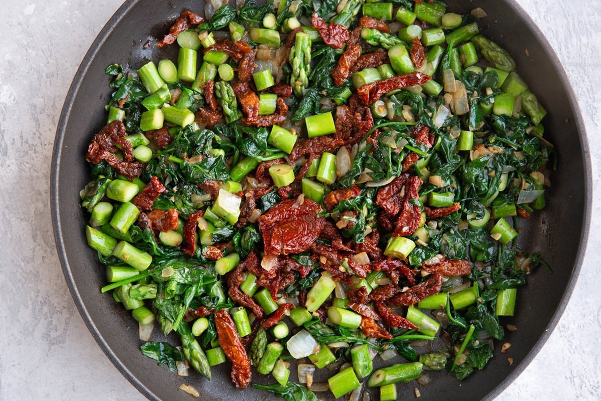 Skillet with sun-dried tomatoes, asparagus, spinach, onion and garlic cooking.