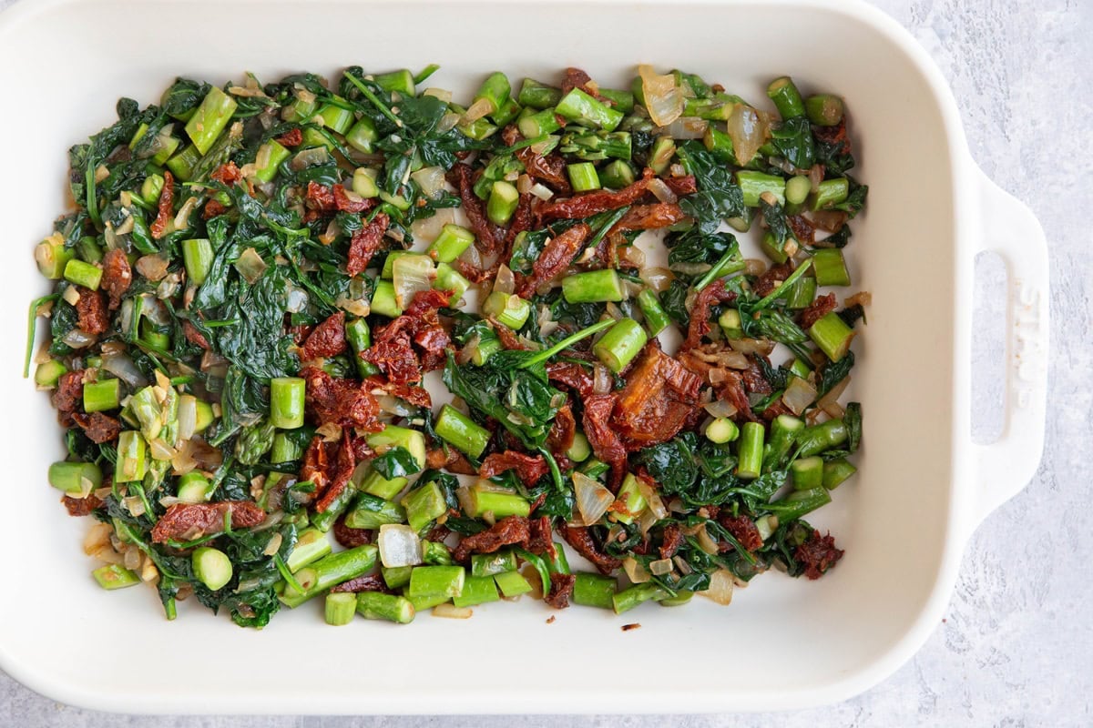 Large casserole dish full of chopped asparagus, spinach, sun-dried tomatoes, onions, and garlic.