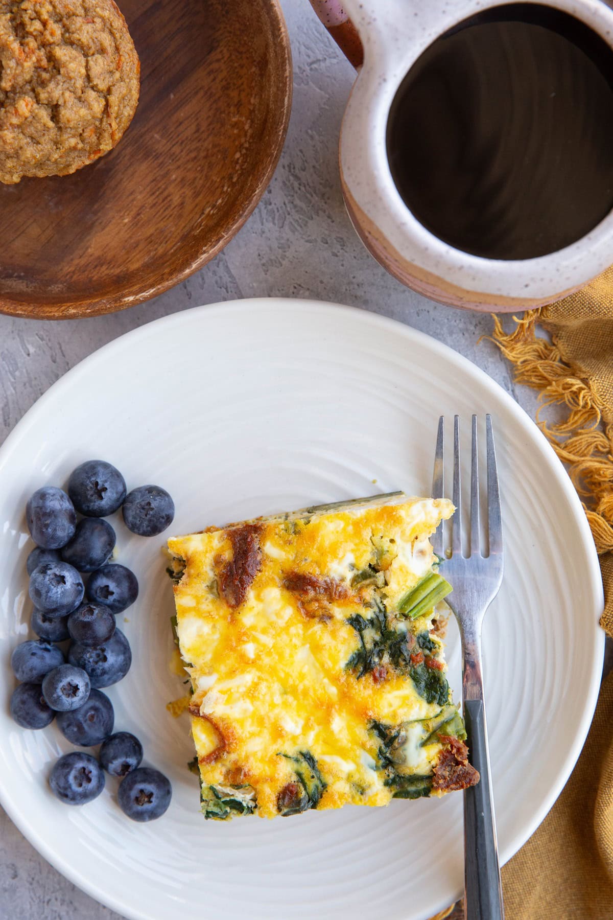 Plate with a slice of asparagus breakfast casserole and fresh blueberries with a cup of coffee and a muffin to the side.