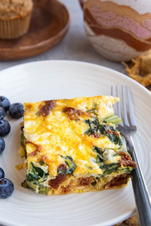 Asparagus and Feta Breakfast Casserole with spinach and sun-dried tomatoes on a white plate with fresh blueberries and a fork to the side.