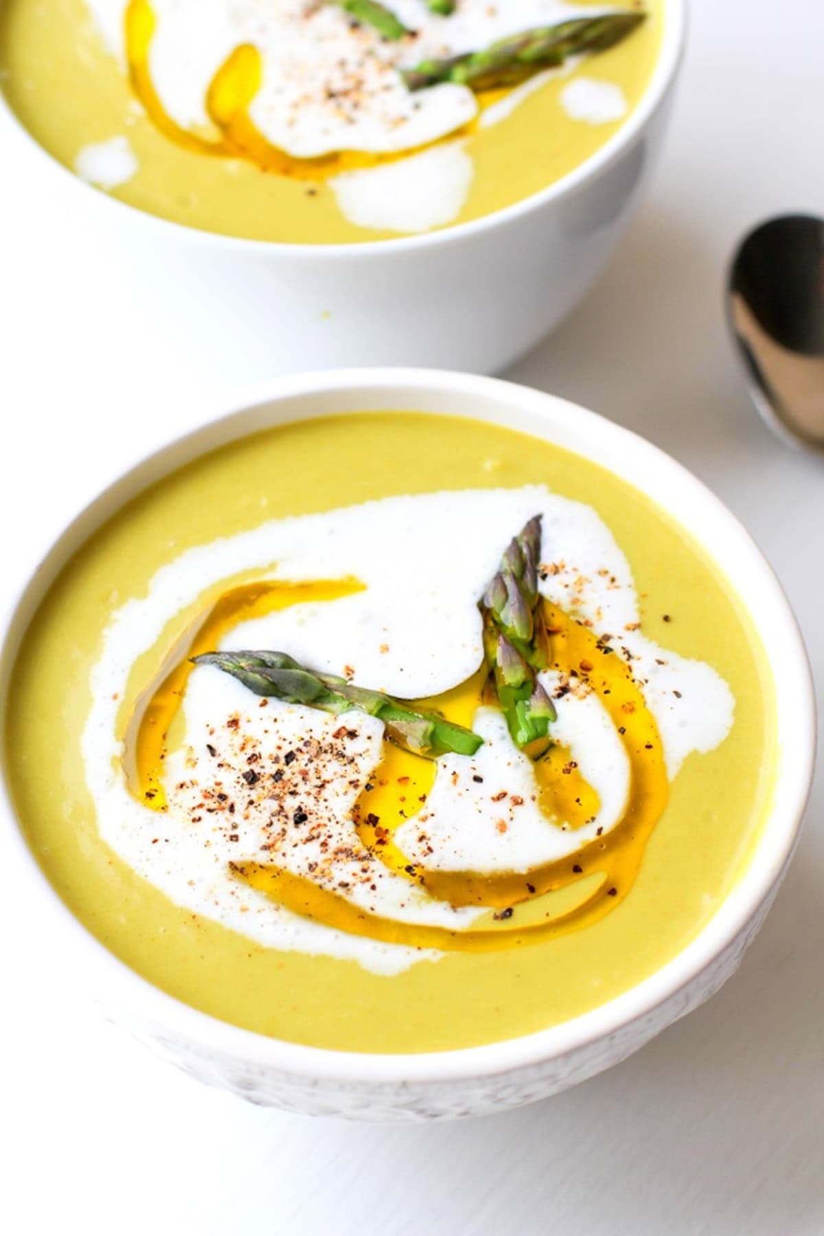 two white bowls full of creamy asparagus soup with asparagus tips floating on top, a drizzle of olive oil, black pepper, and cream.