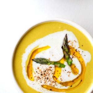 Two white bowls of creamy asparagus soup on a white background with a drizzle of olive oil and heavy cream and cracked pepper on top.