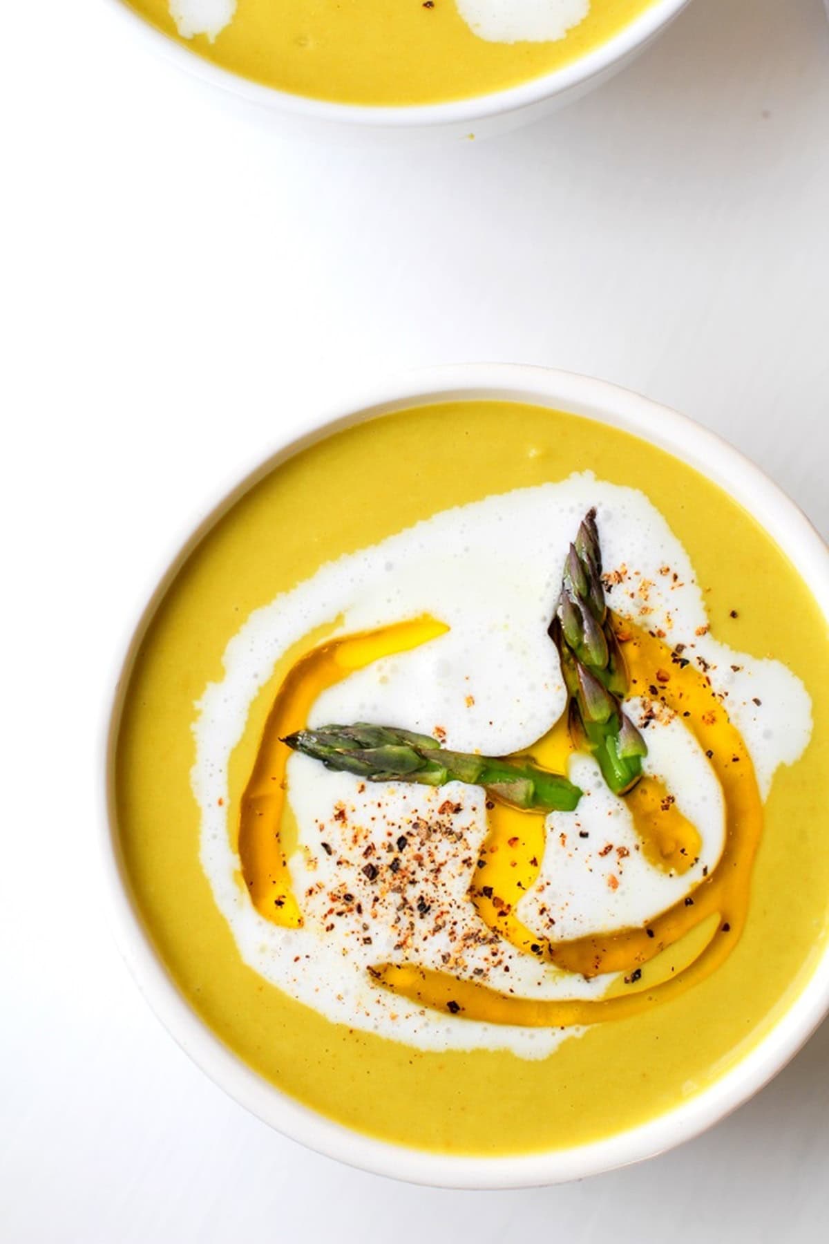 Two white bowls of creamy asparagus soup on a white background with a drizzle of olive oil and heavy cream and cracked pepper on top.
