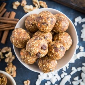 Bowl full of carrot cake energy balls sitting on a dark blue backdrop with flaked coconut, cashews, walnuts, and cinnamon sticks all around.