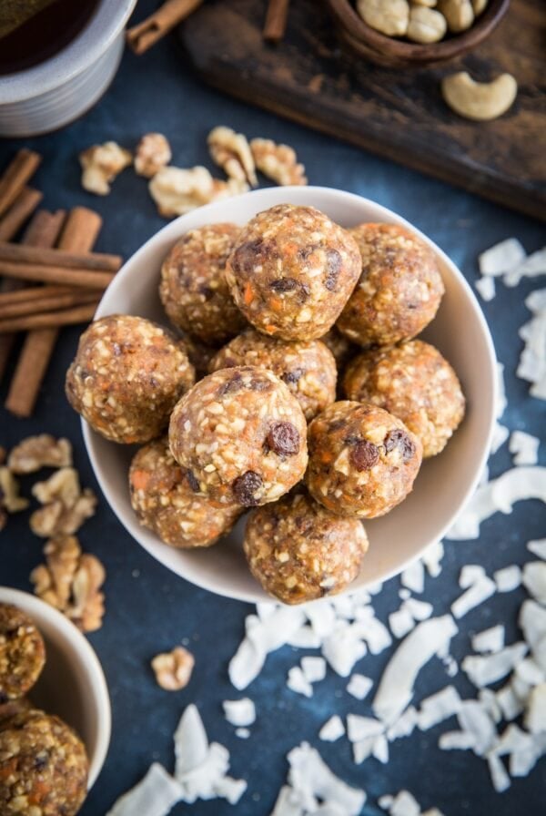 Bowl full of carrot cake energy balls sitting on a dark blue backdrop with flaked coconut, cashews, walnuts, and cinnamon sticks all around.