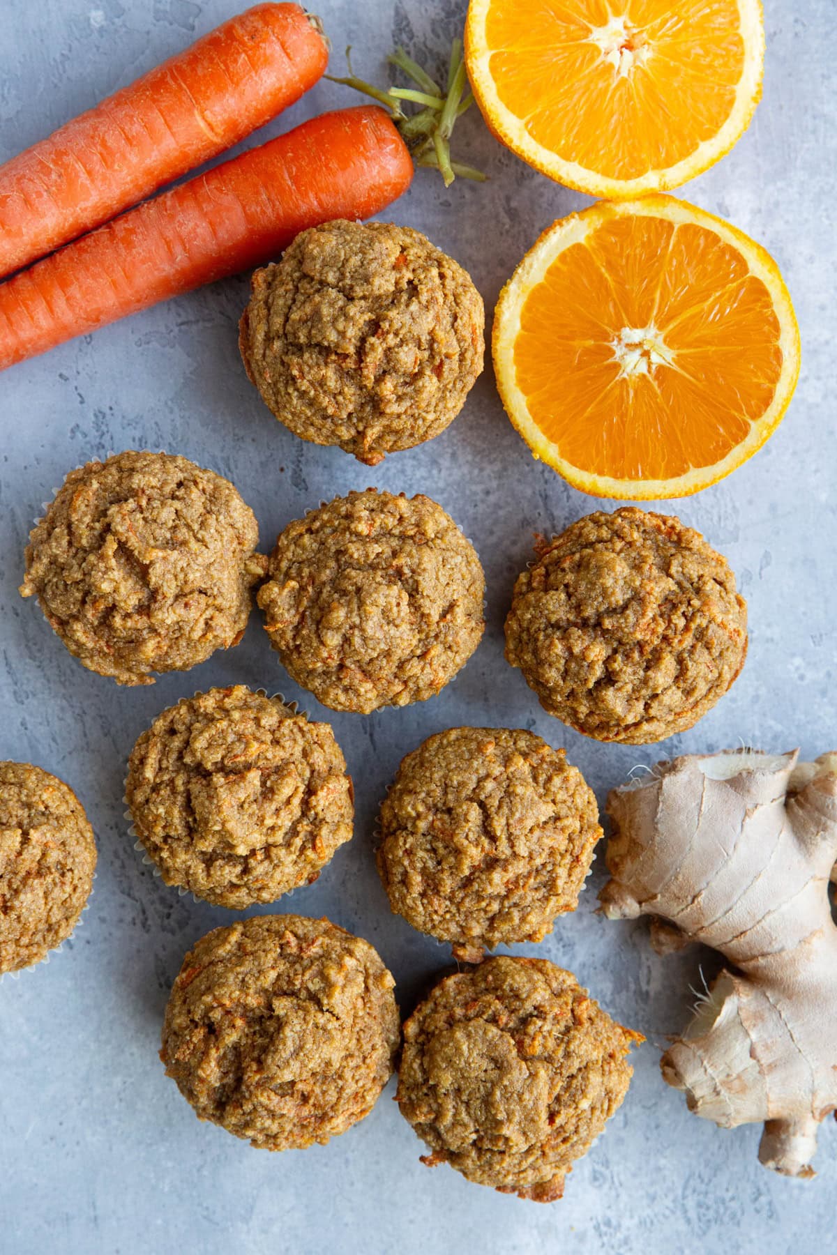 Carrot ginger orange muffins on a backdrop with fresh ginger, carrots, and an orange sliced in half to the side.