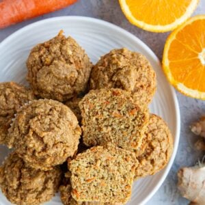 Plate full of carrot orange ginger muffins with one muffin sliced in half so you can see the inside. Fresh ginger, orange, and carrots to the side.