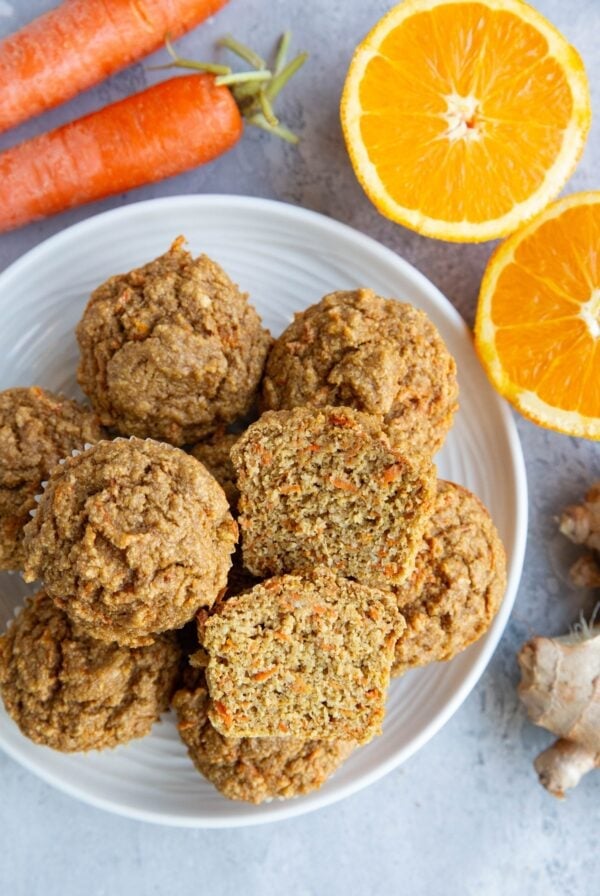 Plate full of carrot orange ginger muffins with one muffin sliced in half so you can see the inside. Fresh ginger, orange, and carrots to the side.