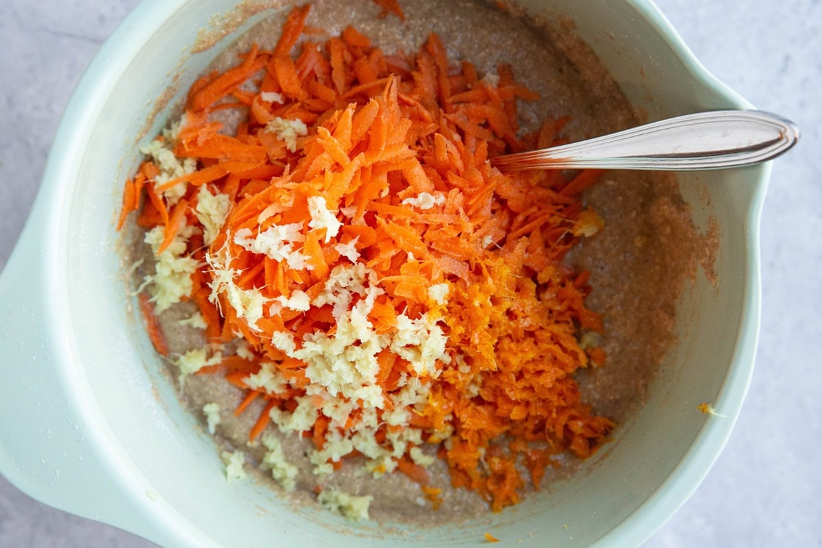 Mixing bowl with bread batter on the bottom and grated carrots, ginger, and orange zest on top, ready to be mixed in.
