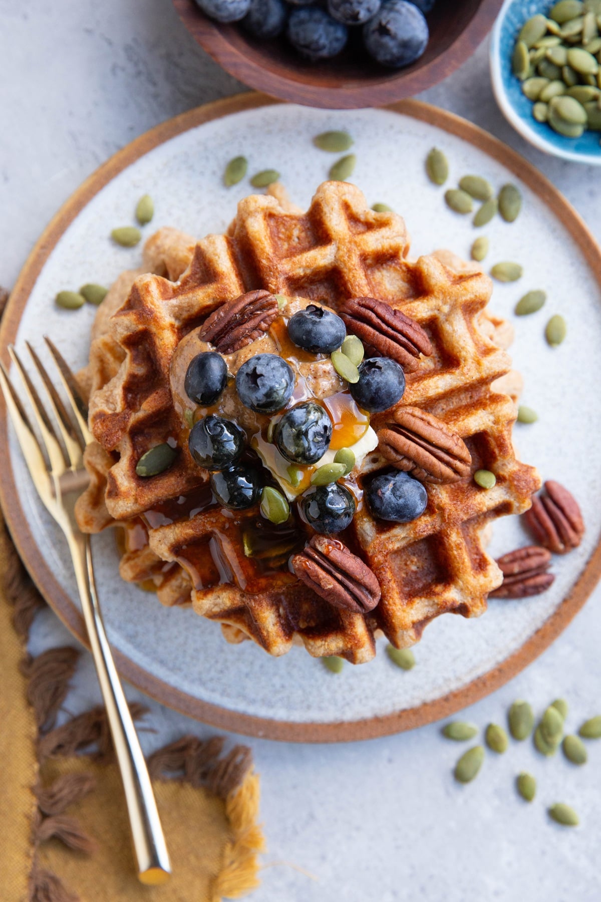 Plate of waffles with almond butter, regular butter, blueberries, pumpkin seeds and pecans on top. A gold fork to the side and bowls of blueberries and pumpkin seeds.