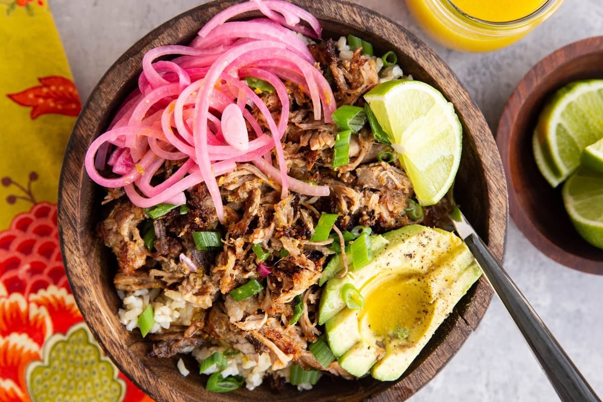 Large wooden bowl full of crispy pork on top of brown rice with sliced avocado and pickled red onions.