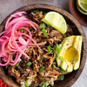 Big wooden bowl full of brown rice, shredded pork, pickled red onions, sliced avocado and a slice of lime, ready to serve.
