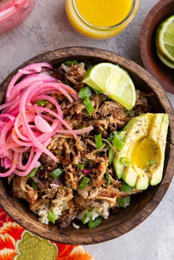 Big wooden bowl full of brown rice, shredded pork, pickled red onions, sliced avocado and a slice of lime, ready to serve.