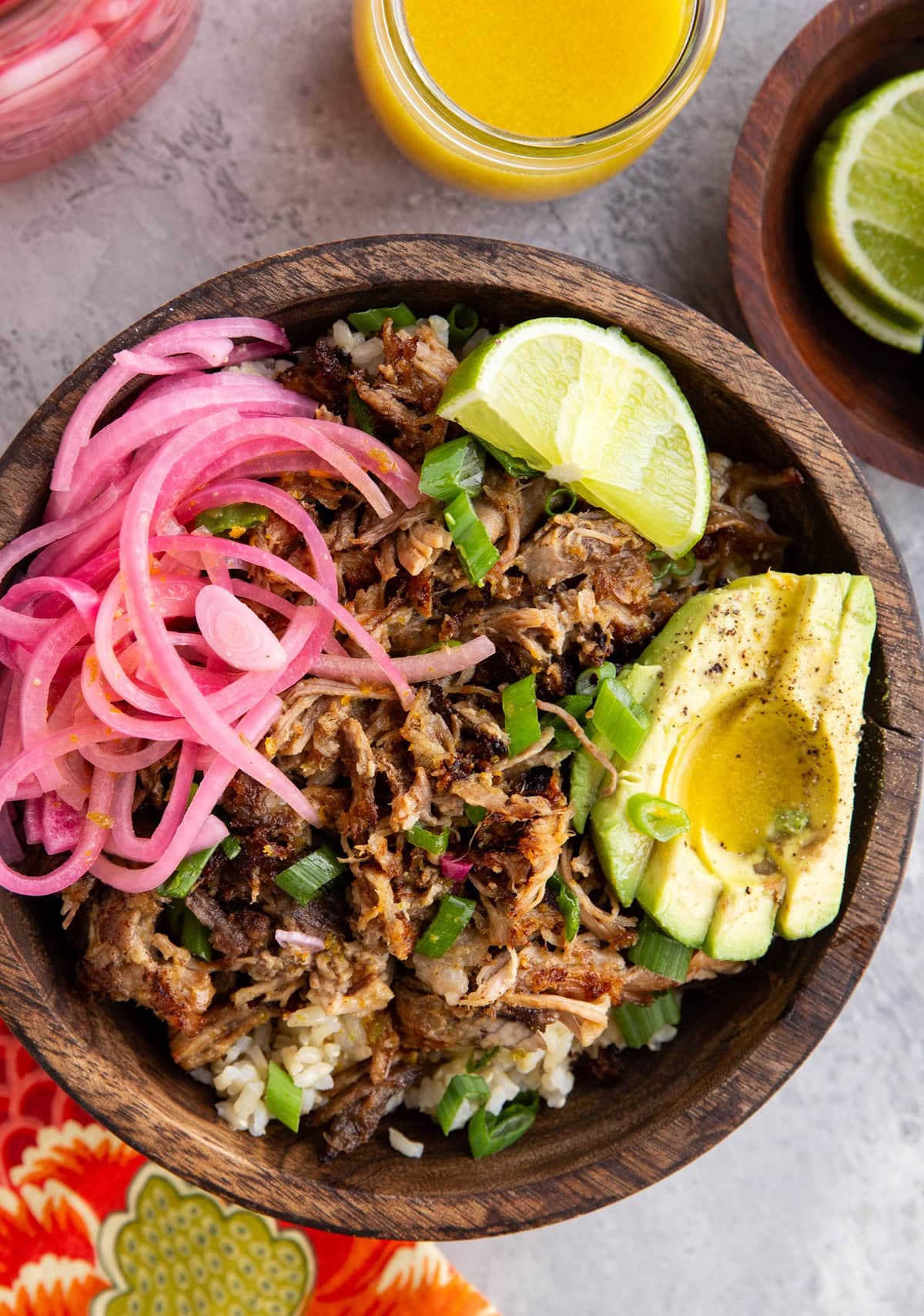 Big wooden bowl full of brown rice, shredded pork, pickled red onions, sliced avocado and a slice of lime, ready to serve.