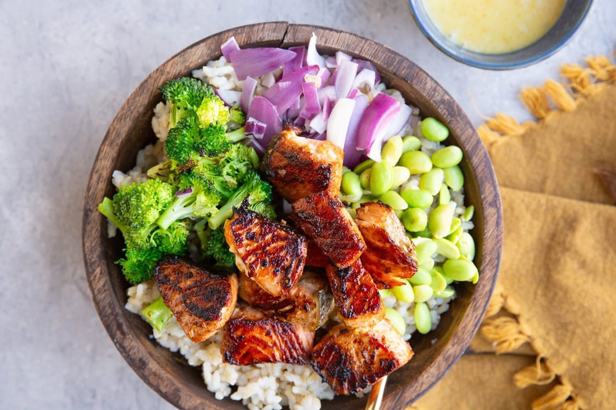 Wooden bowl packed with brown rice, seared salmon bites, broccoli, red onion, edamame and a bowl of mojo sauce to the side.