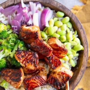 Seared salmon bites in a wooden bowl with chopped red onions, broccoli, and edamame with a bowl of mojo sauce to the side.