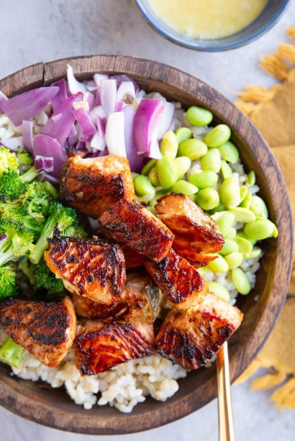 Seared salmon bites in a wooden bowl with chopped red onions, broccoli, and edamame with a bowl of mojo sauce to the side.