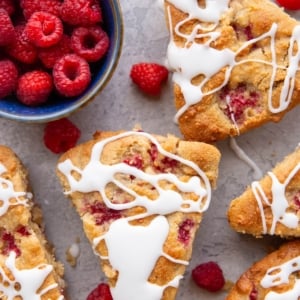 Six raspberry scones sitting on a background drizzled with glaze with fresh raspberries all around and a small bowl of raspberries to the side.
