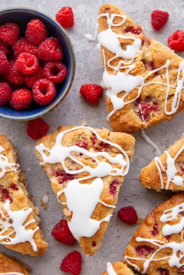 Six raspberry scones sitting on a background drizzled with glaze with fresh raspberries all around and a small bowl of raspberries to the side.