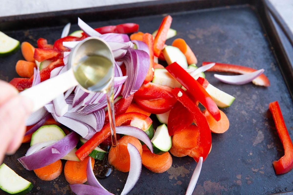 Hand pouring avocado oil over chopped vegetables on a sheet pan.