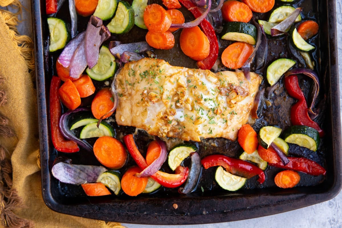 Sheet pan with vegetables and one cooked cod fillet, fresh out of the oven and ready to serve.